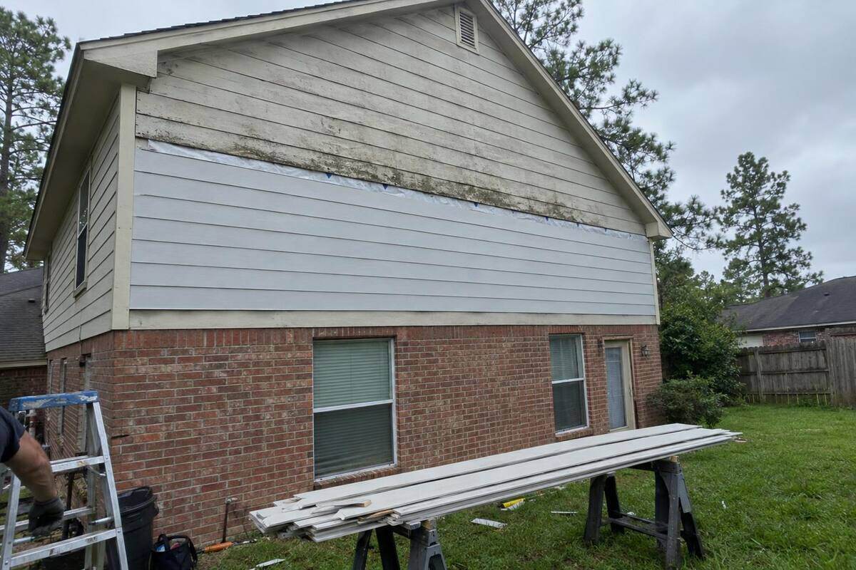 Upper gable wall with new lap planks below and old swollen siding above, planks on sawhorses on the lawn