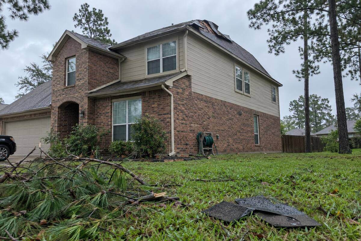 Two-story brick house with shingles folded back at the ridge and loose shingles in the wet grass